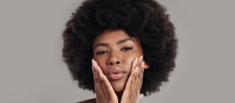 Mulher negra jovem com cabelo crespo estilizado em um black power posando para uma foto com as duas mãos posicionadas ao lado do rosto.