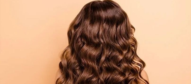 Mulher com cabelo castanho e ondulado longo de costas para a câmera, mostrando sua finalização capilar definida.