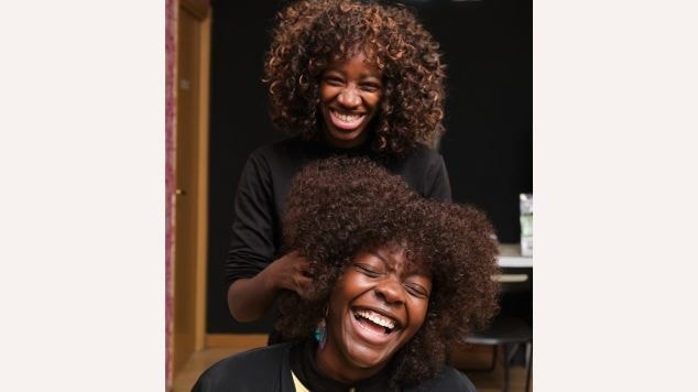 Profissional faz o corte de franja no cabelo crespo em uma mulher negra e sorridente