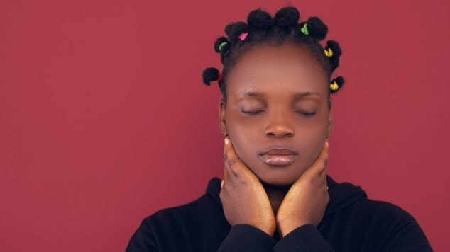 Mulher de olhos fechados com moletom preto e cabelo com penteado bantu knots apoiando o queixo nas mãos que estão envolvendo o pescoço.