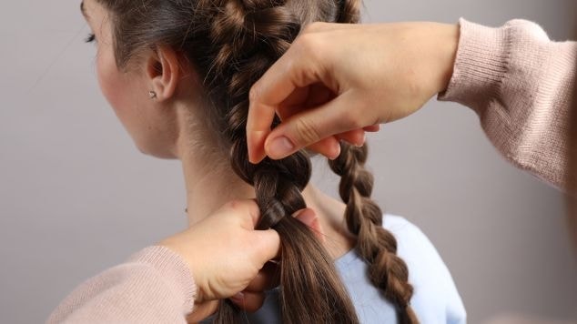 Mãos trançando cabelo castanho em duas maria-chiquinhas, close no processo de finalização das tranças na altura da nuca.