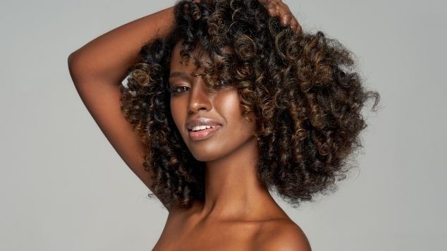 Mulher negra sorrindo, exibindo cachos volumosos e definidos em tons castanhos, com uma mão erguida tocando os fios cheios de textura natural.